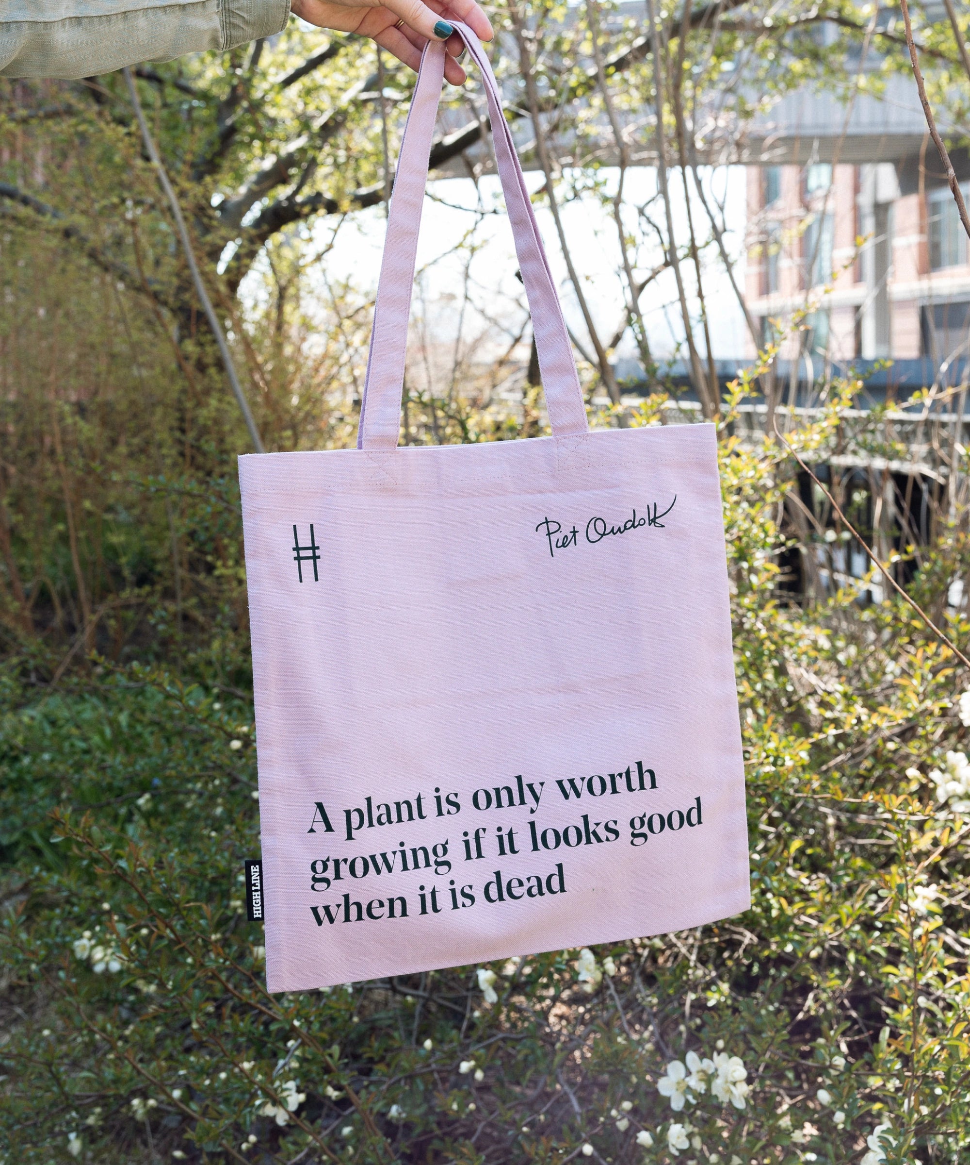 Piet Oudolf Quote Tote in Aster—a pale purple cotton canvas tote bag featuring the quote "A plant is only worth growing if it looks good when it is dead." The tote is being held up against the foliage of the High Line.