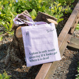 Piet Oudolf Quote Tote in Aster—a pale purple cotton canvas tote bag featuring the quote "A plant is only worth growing if it looks good when it is dead." Tote is shown leaning against a pruned tree and the rail tracks on the High Line.