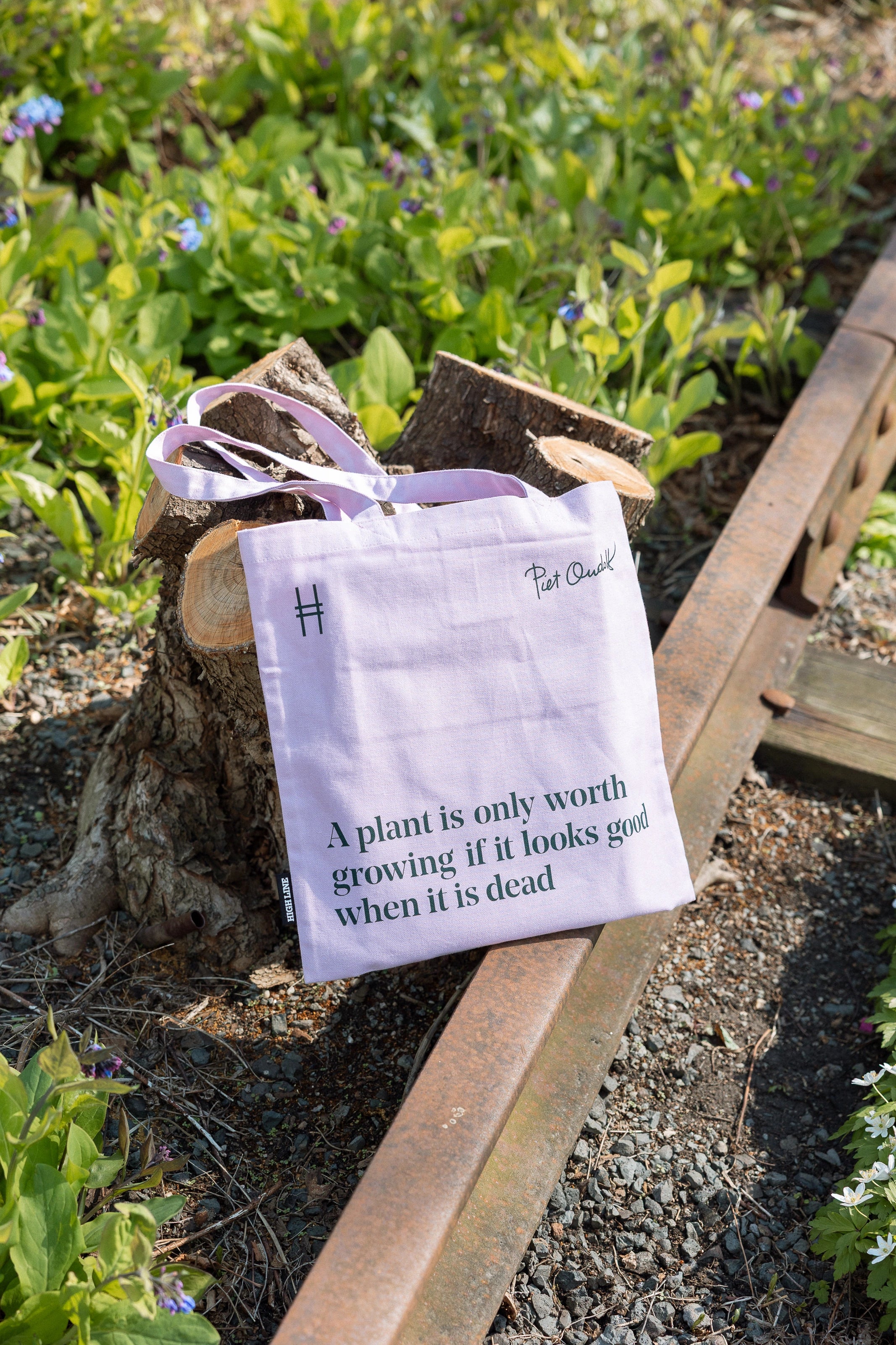 Piet Oudolf Quote Tote in Aster—a pale purple cotton canvas tote bag featuring the quote "A plant is only worth growing if it looks good when it is dead." Tote is shown leaning against a pruned tree and the rail tracks on the High Line.