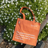 Piet Oudolf Quote Tote in Trumpet Honeysuckle—an orange cotton canvas tote bag featuring the quote "The garden is a promise. And I think that promise is why you want to come back." The tote is resting on the High Line rail tracks against a flowering shrub.