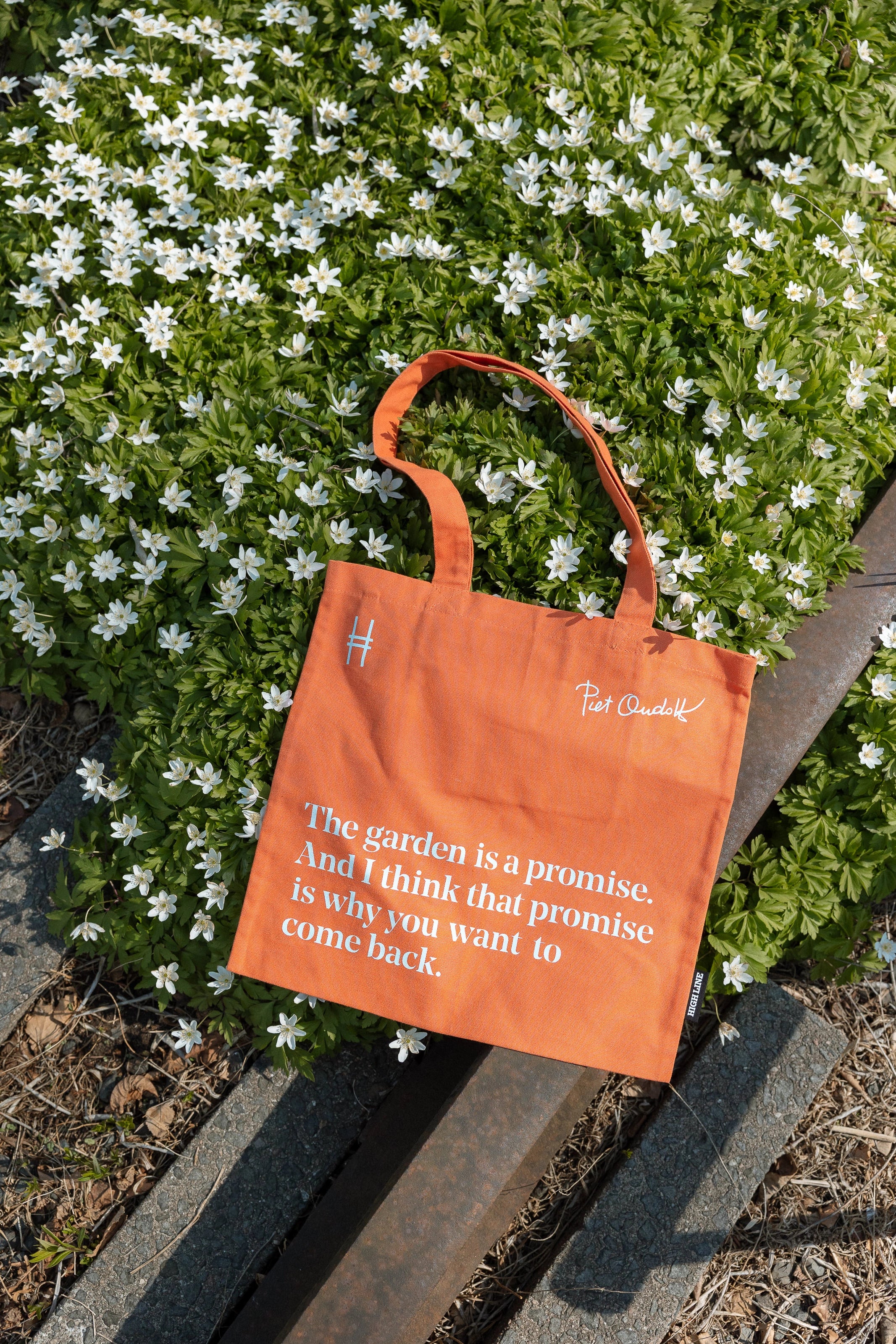 Piet Oudolf Quote Tote in Trumpet Honeysuckle—an orange cotton canvas tote bag featuring the quote "The garden is a promise. And I think that promise is why you want to come back." The tote is resting on the High Line rail tracks against a flowering shrub.