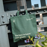 Piet Oudolf quote tote in Ivy, featuring the quote "You have to know your plants" hanging off of the rail of the High Line, flowers around it.
