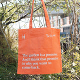 Piet Oudolf Quote Tote in Trumpet Honeysuckle—an orange cotton canvas tote bag featuring the quote "The garden is a promise. And I think that promise is why you want to come back." The bag is being held up against the foliage of the High Line.