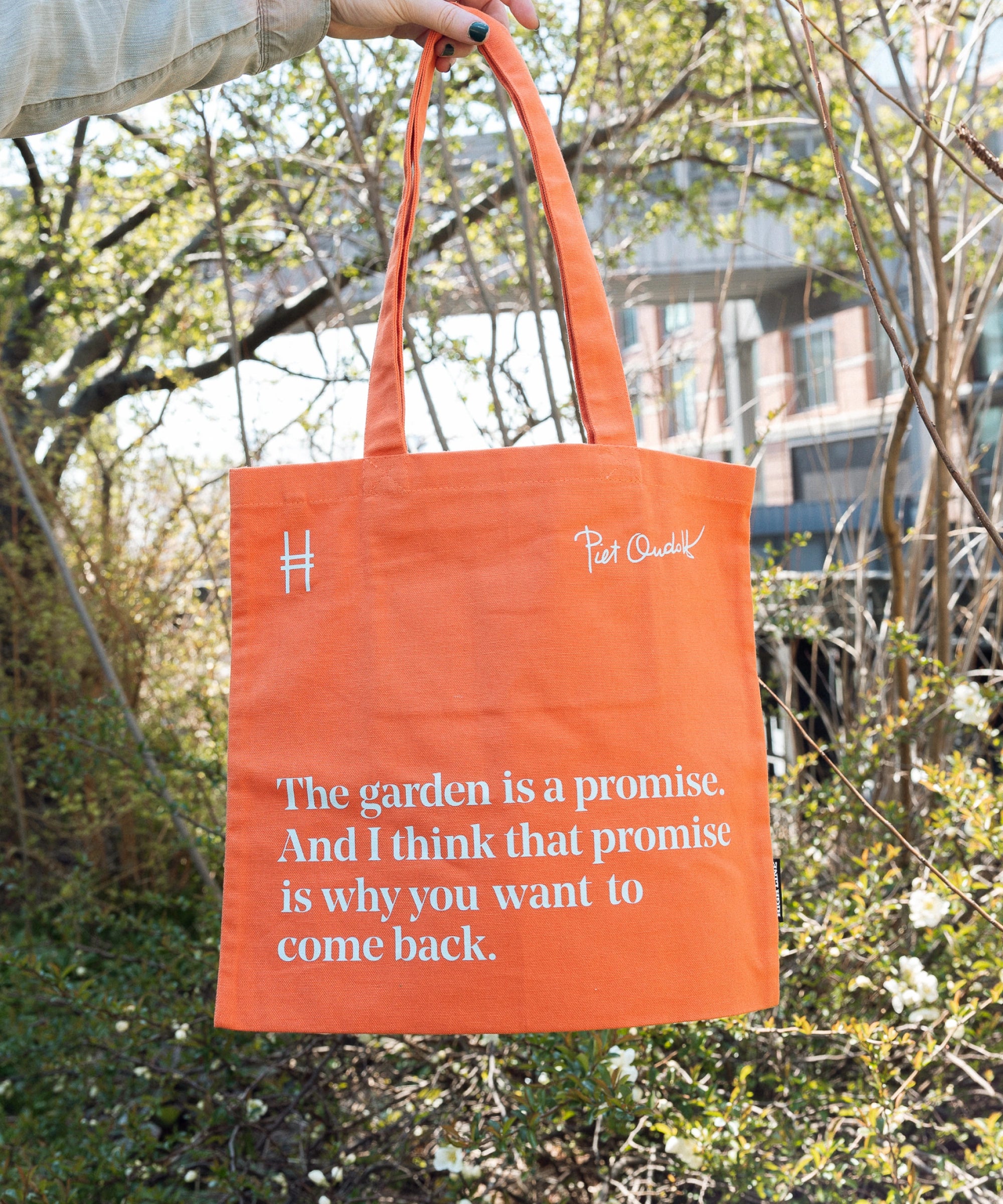 Piet Oudolf Quote Tote in Trumpet Honeysuckle—an orange cotton canvas tote bag featuring the quote "The garden is a promise. And I think that promise is why you want to come back." The bag is being held up against the foliage of the High Line.