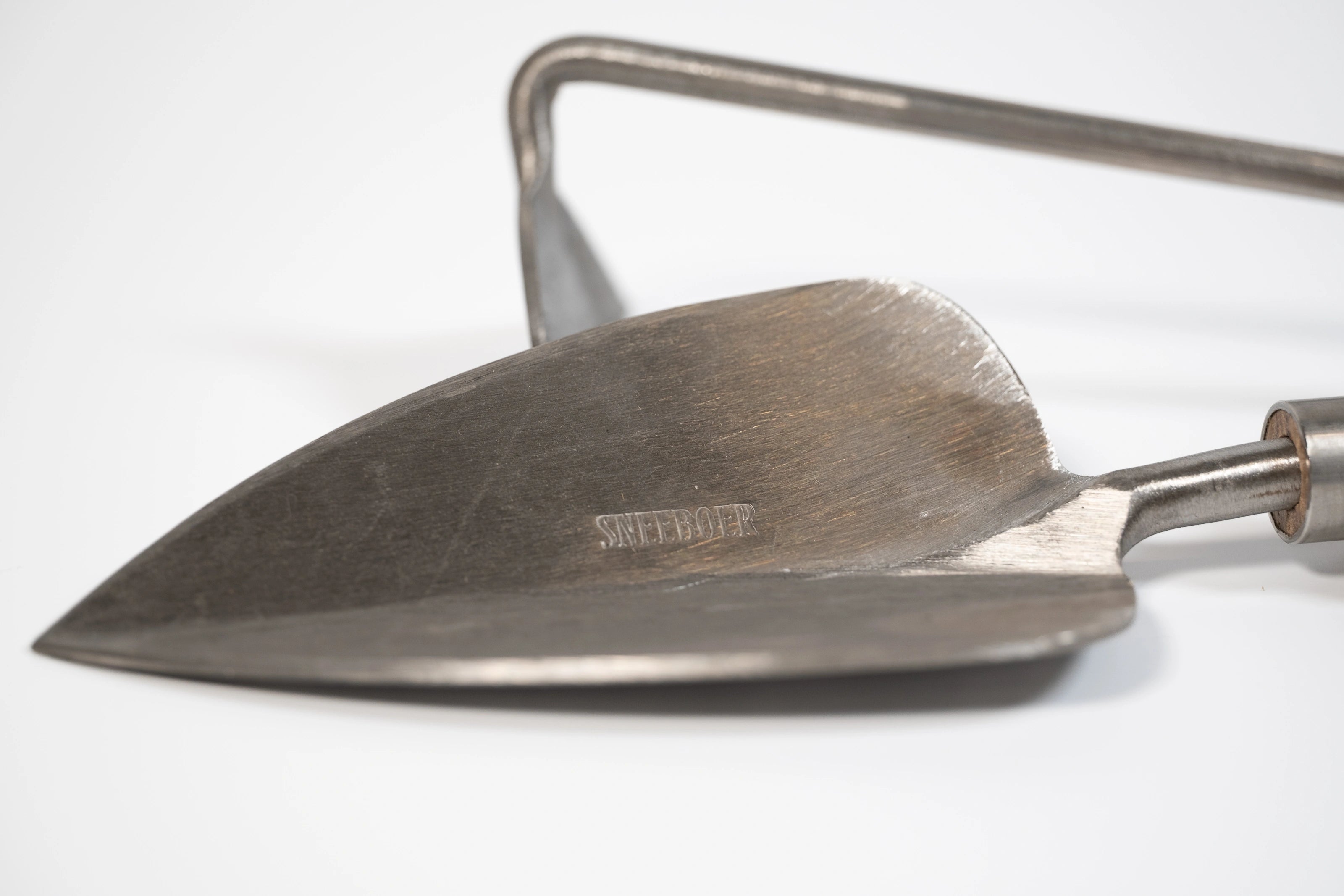 Closeup on the stainless steel trowel blade with hand hoe in the background. Sneeboer logo embossed on the trowel blade.