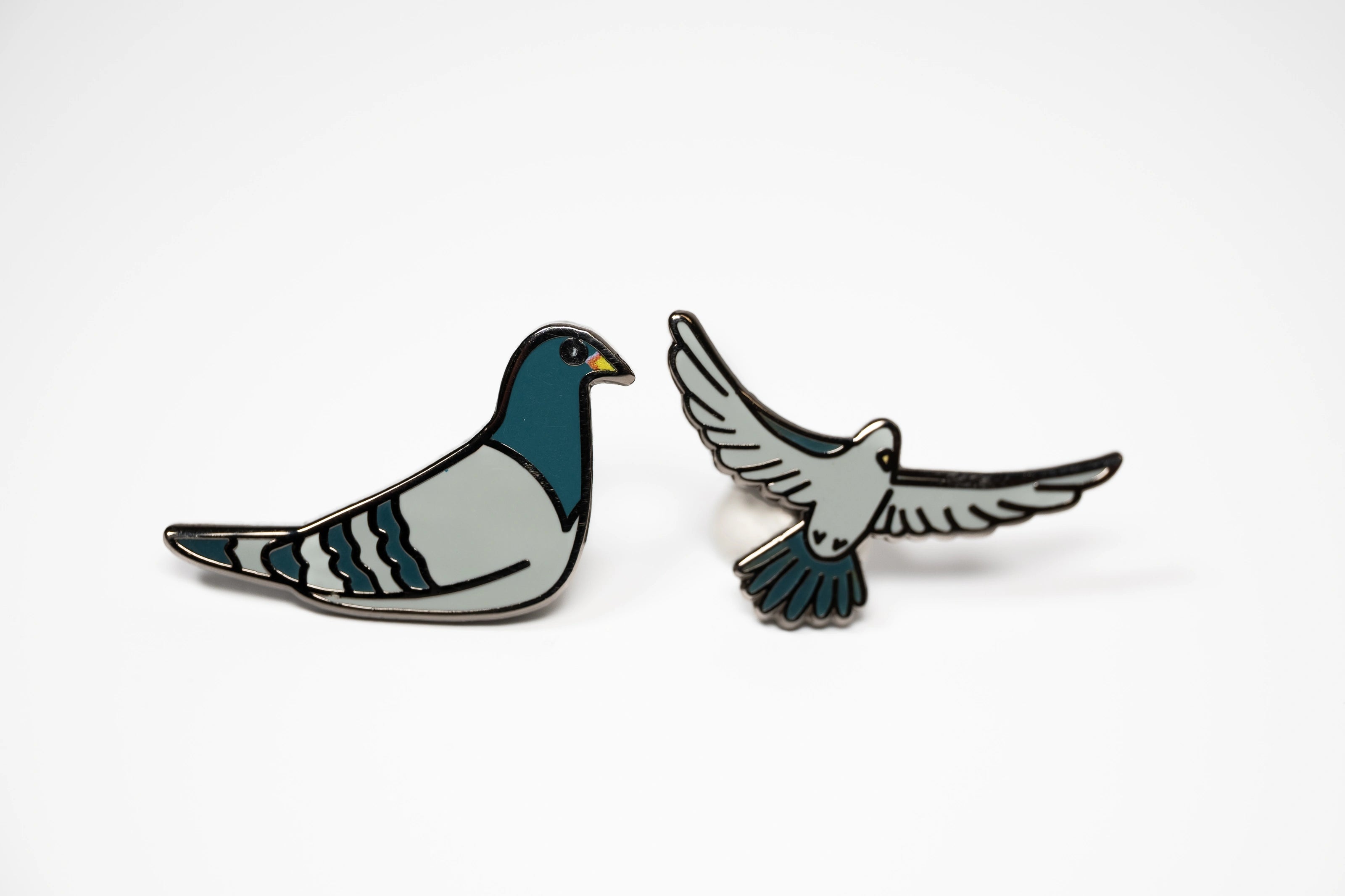 The Pigeon Pins—two enamel pigeon pins, one sitting and one in flight—on a white background.