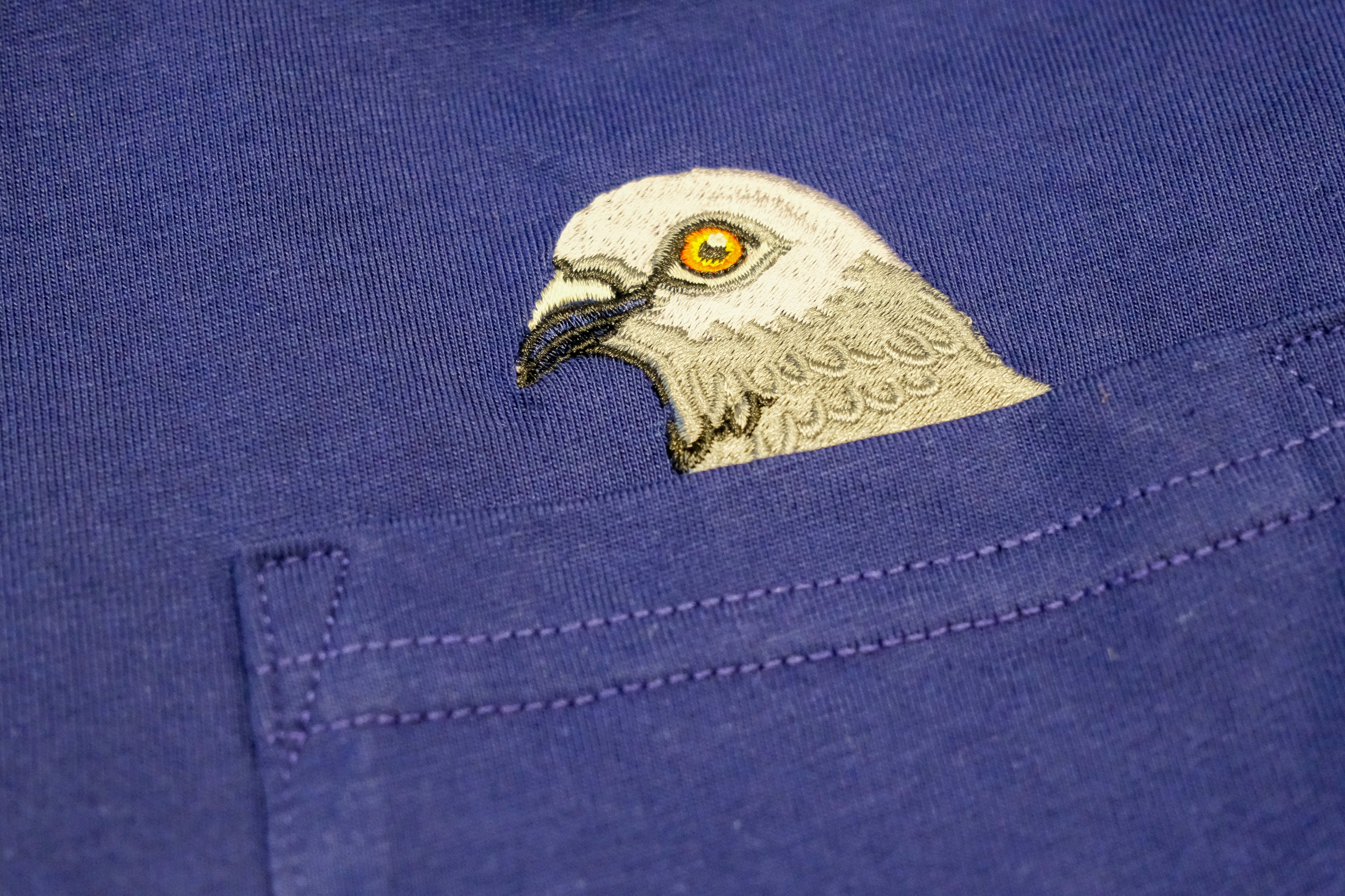 Closeup of Pigeon head embroidery popping out of the pocket on the Pigeon Pocket long-sleeve tee in Navy a rich, saturated, dark blue.