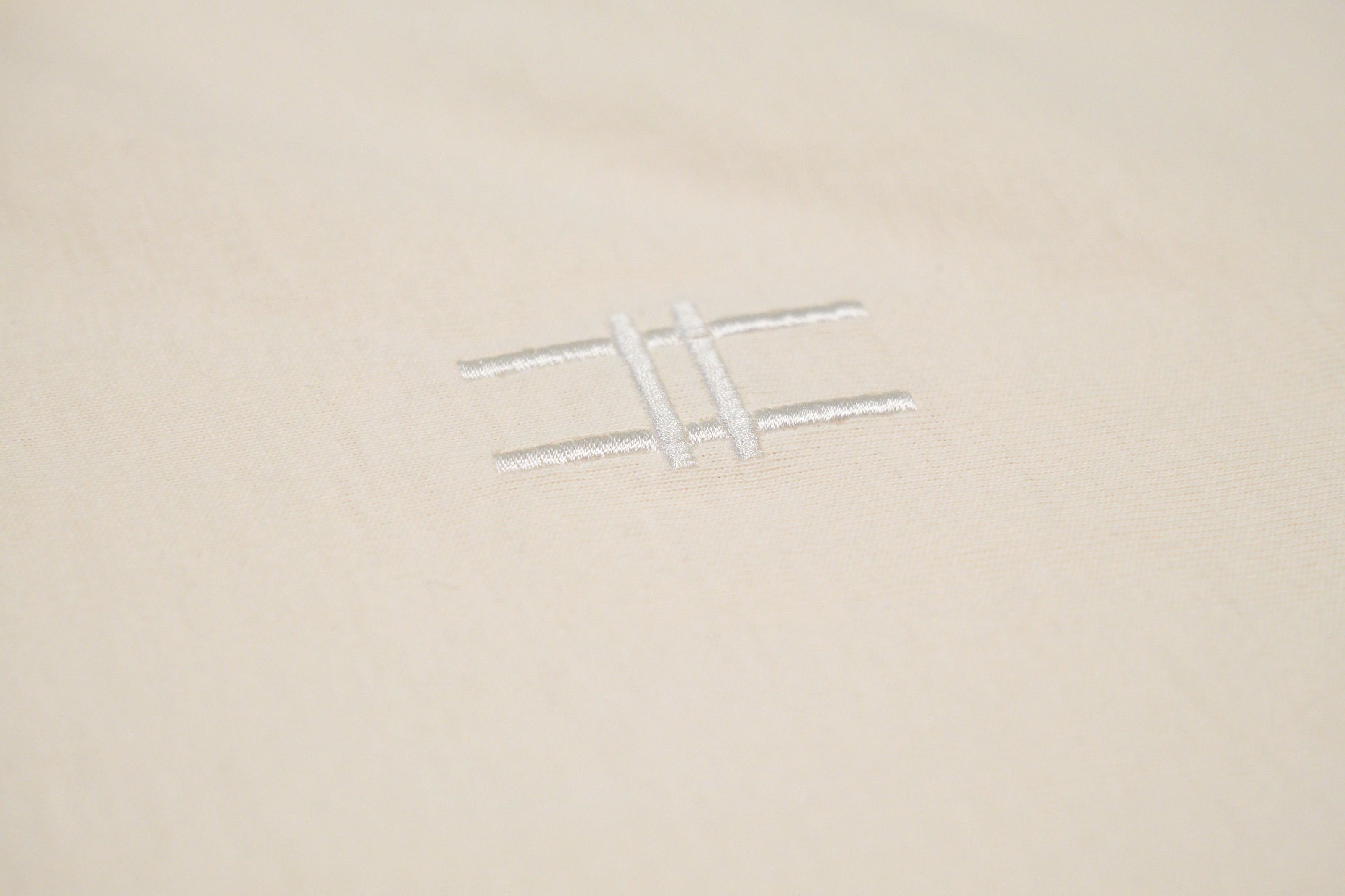 Closeup of the High Line icon, a railroad-track-inspired "H" which is embroidered in off-white thread to match the natural, 100% cotton fabric of the Pigeon Pocket long-sleeve tee in River Birch.