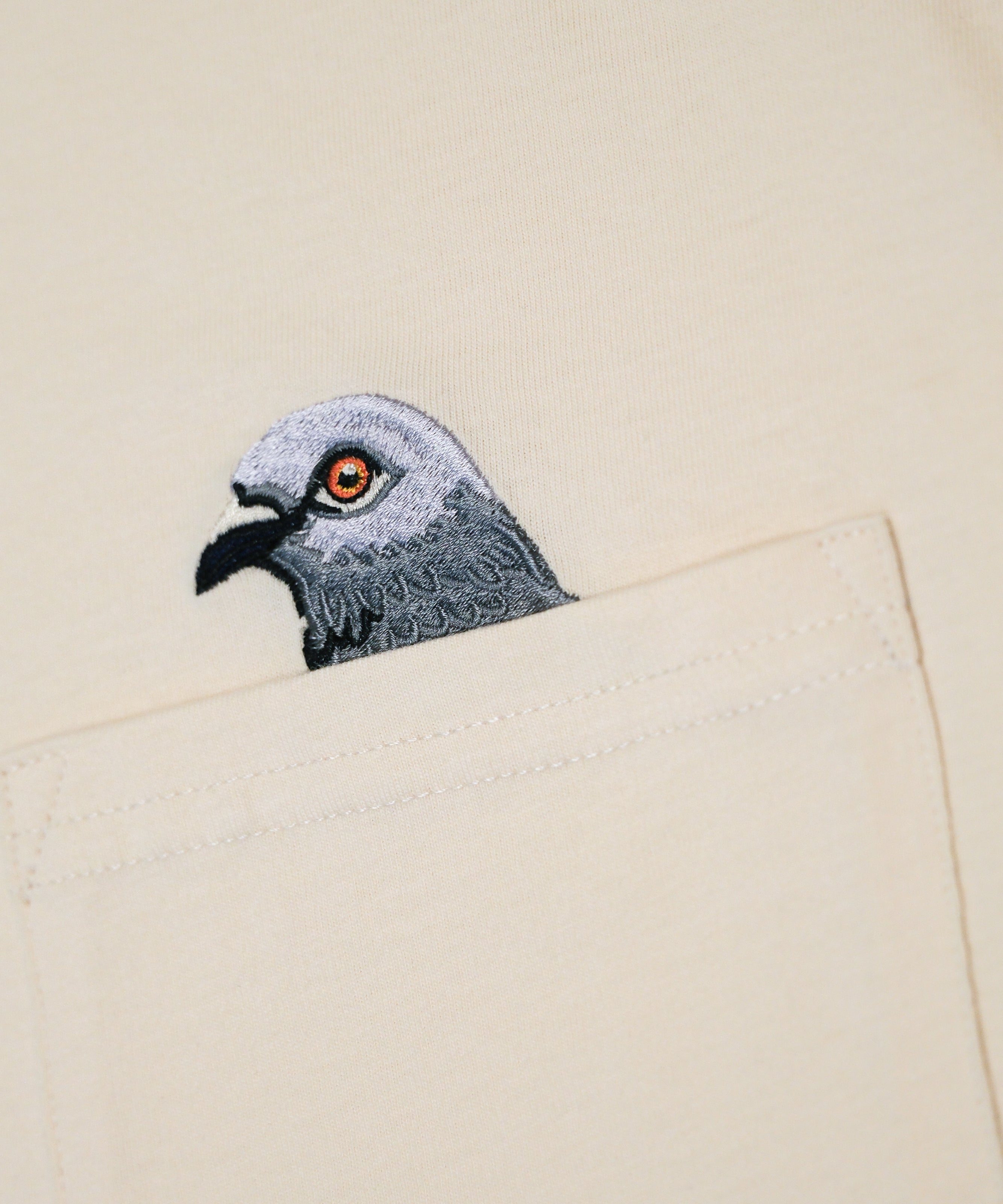Closeup of Pigeon head embroidery popping out of the pocket on the Pigeon Pocket long-sleeve tee in River Birch, a natural off-white color.