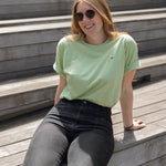 Woman wearing a light green crewneck t-shirt with a small screen-printed hummingbird on the left chest, paired with dark jeans, sits on the Sunken Overlook steps on the High Line.