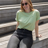 Woman wearing a light green crewneck t-shirt with a small screen-printed hummingbird on the left chest, paired with dark jeans, sits on the Sunken Overlook steps on the High Line.