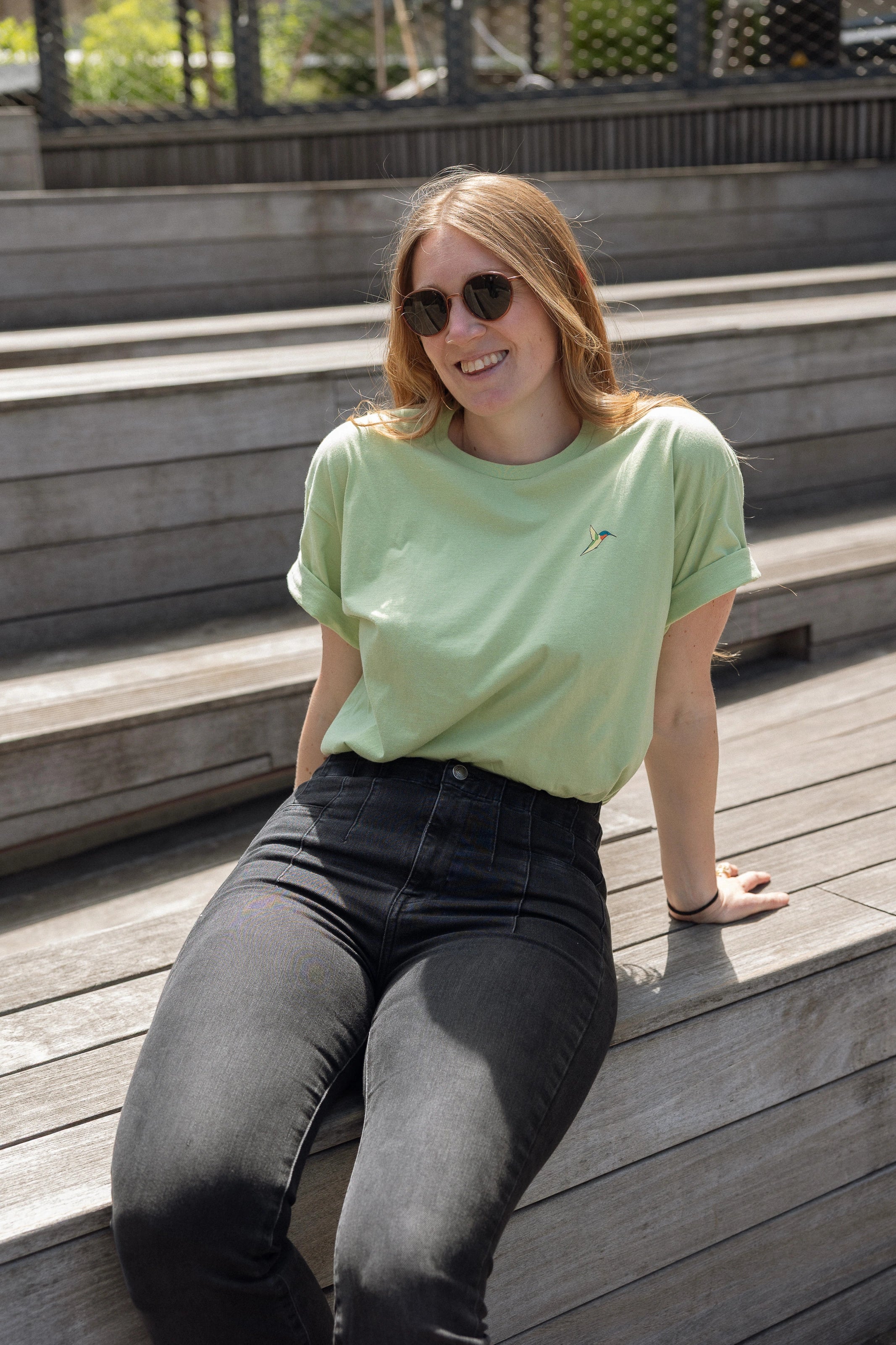 Woman wearing a light green crewneck t-shirt with a small screen-printed hummingbird on the left chest, paired with dark jeans, sits on the Sunken Overlook steps on the High Line.