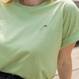 A close-up of a person wearing a light green crewneck t-shirt with a small screen-printed hummingbird on the left chest, paired with dark jeans.