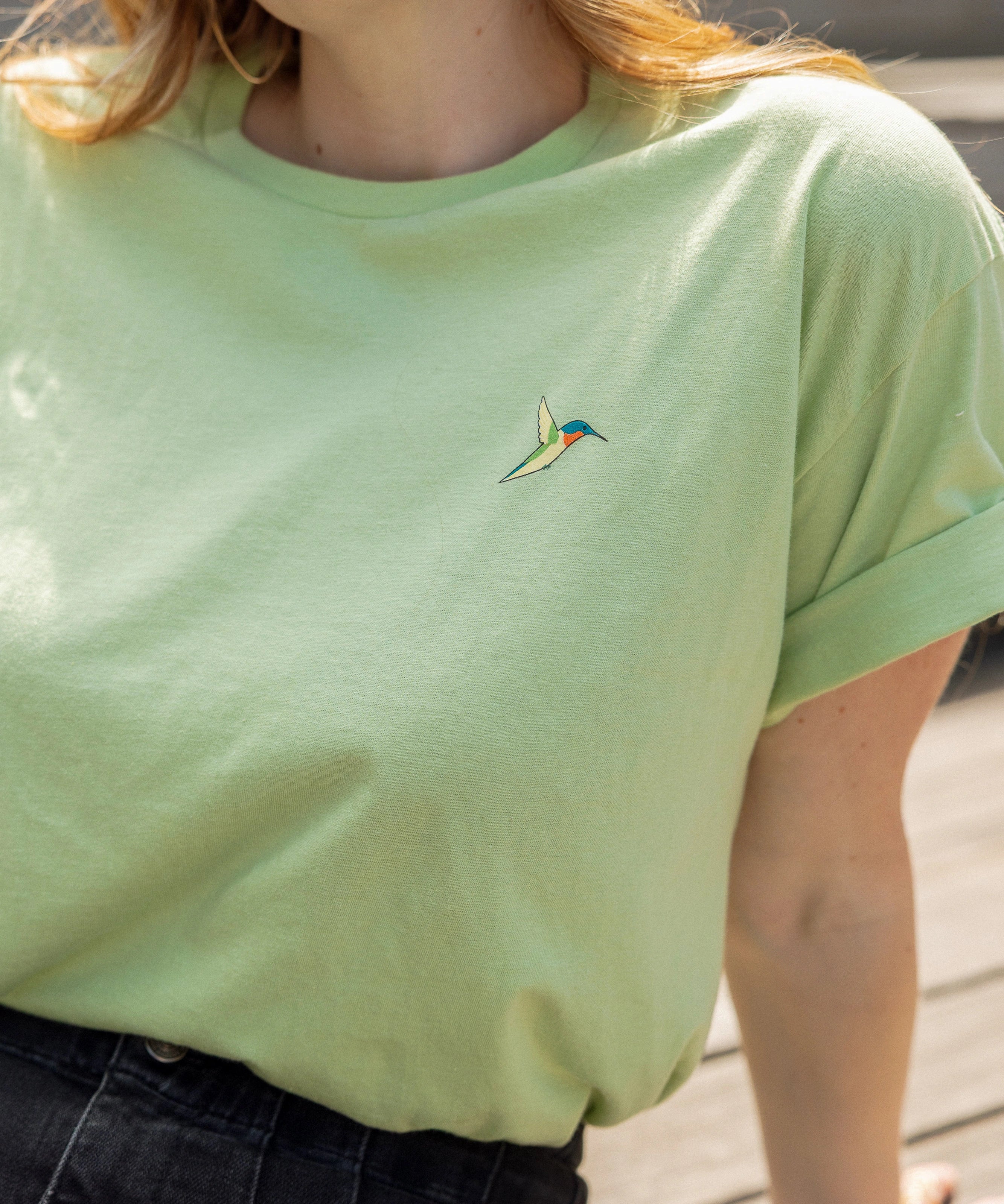 A close-up of a person wearing a light green crewneck t-shirt with a small screen-printed hummingbird on the left chest, paired with dark jeans.