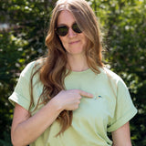 A woman wearing a light green crewneck t-shirt with a small screen-printed hummingbird on the left chest, which she's pointing to. She is wearing dark sunglasses and dark jeans, and is amongst the foliage on the High Line.