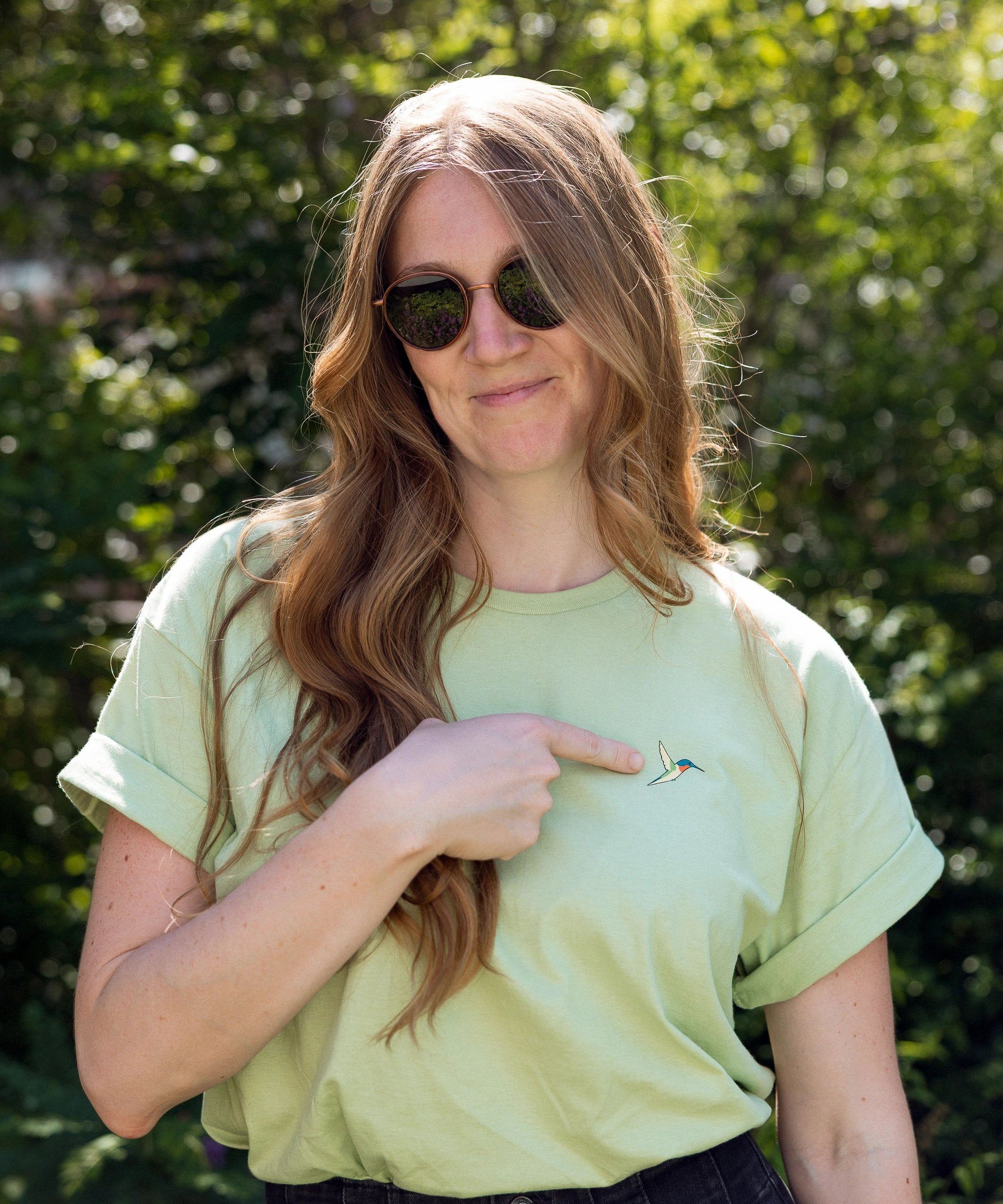 A woman wearing a light green crewneck t-shirt with a small screen-printed hummingbird on the left chest, which she's pointing to. She is wearing dark sunglasses and dark jeans, and is amongst the foliage on the High Line.