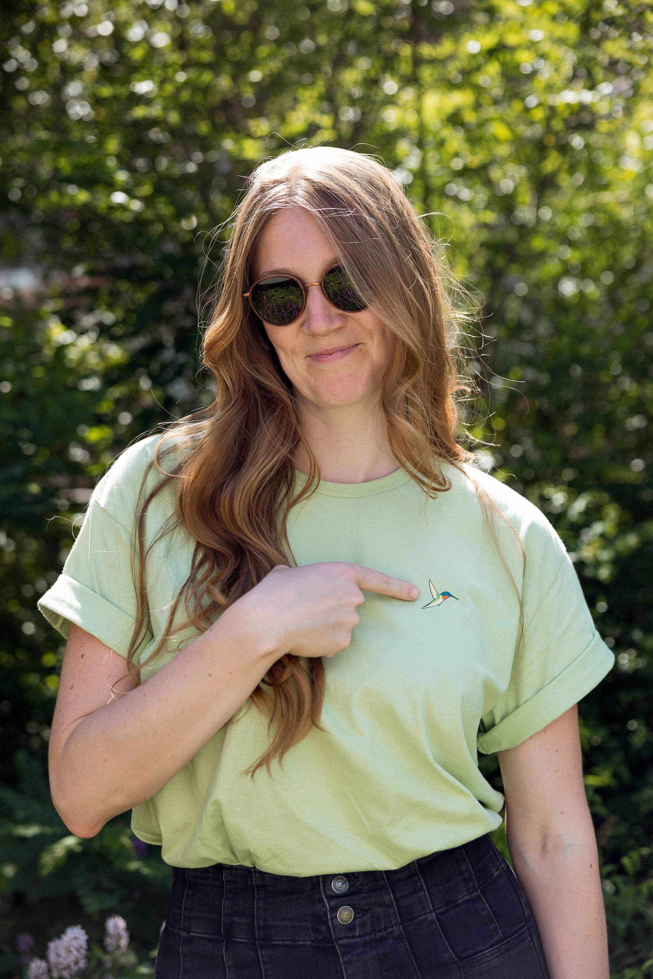 A woman wearing a light green crewneck t-shirt with a small screen-printed hummingbird on the left chest, which she's pointing to. She is wearing dark sunglasses and dark jeans, and is amongst the foliage on the High Line.