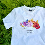 White t-shirt featuring a flower crown and text reading 'FOR ALL OF US' on a bed of ferns.