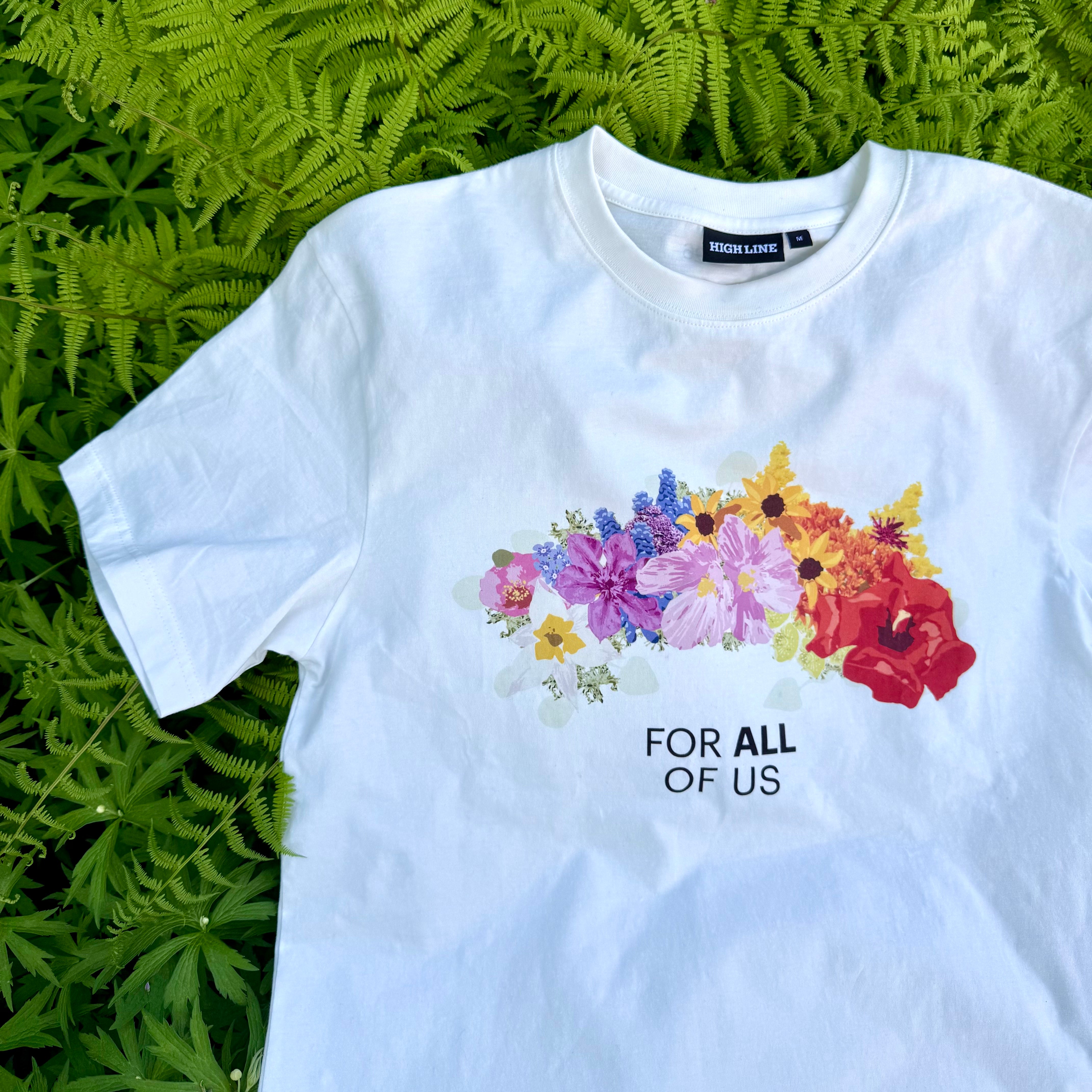 White t-shirt featuring a flower crown and text reading 'FOR ALL OF US' on a bed of ferns.