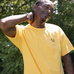 A man wearing a bright yellow crewneck t-shirt with a small bumblebee screenprinted on the left chest, posing outdoors against the foliage of the High Line.