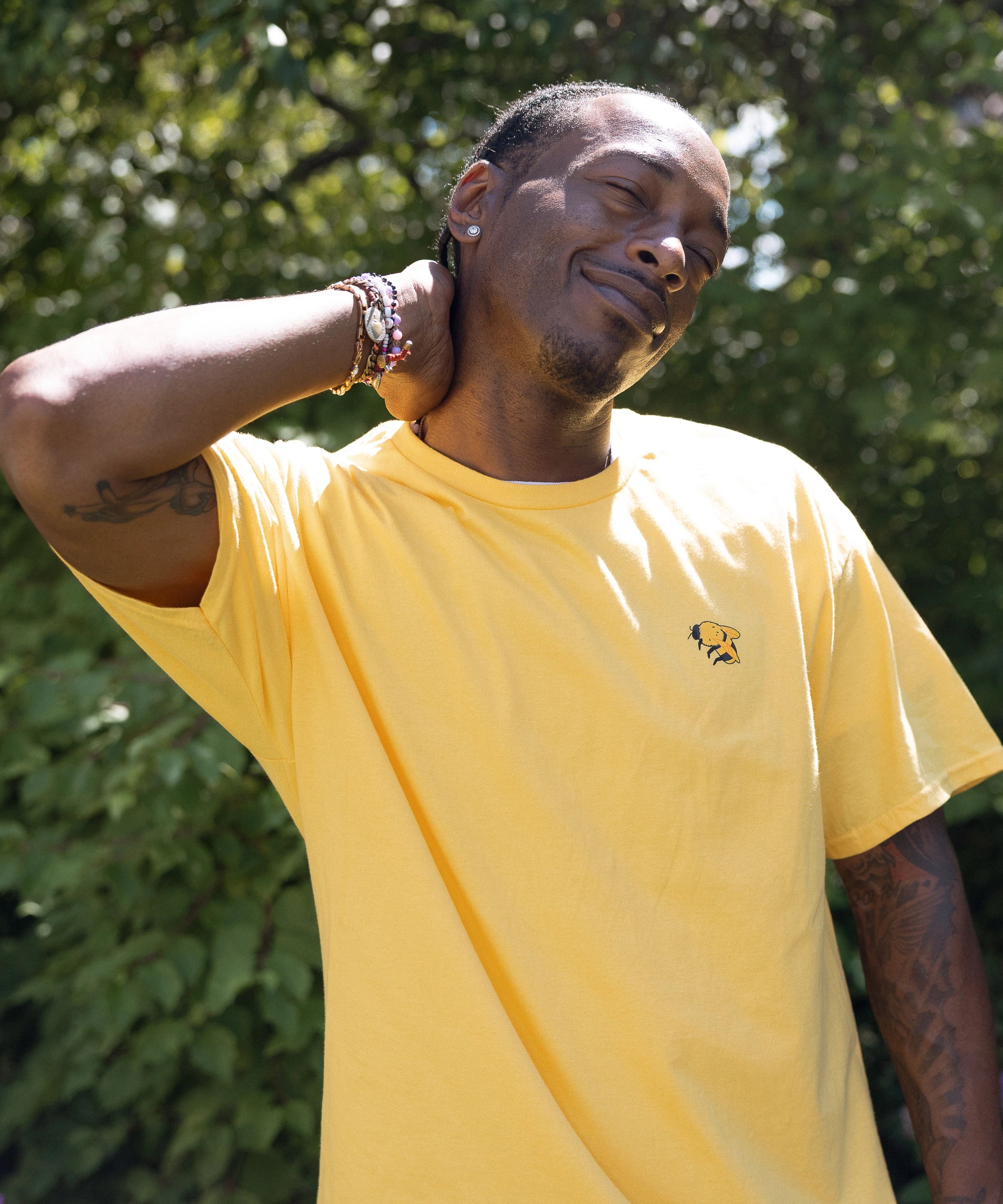 A man wearing a bright yellow crewneck t-shirt with a small bumblebee screenprinted on the left chest, posing outdoors against the foliage of the High Line.