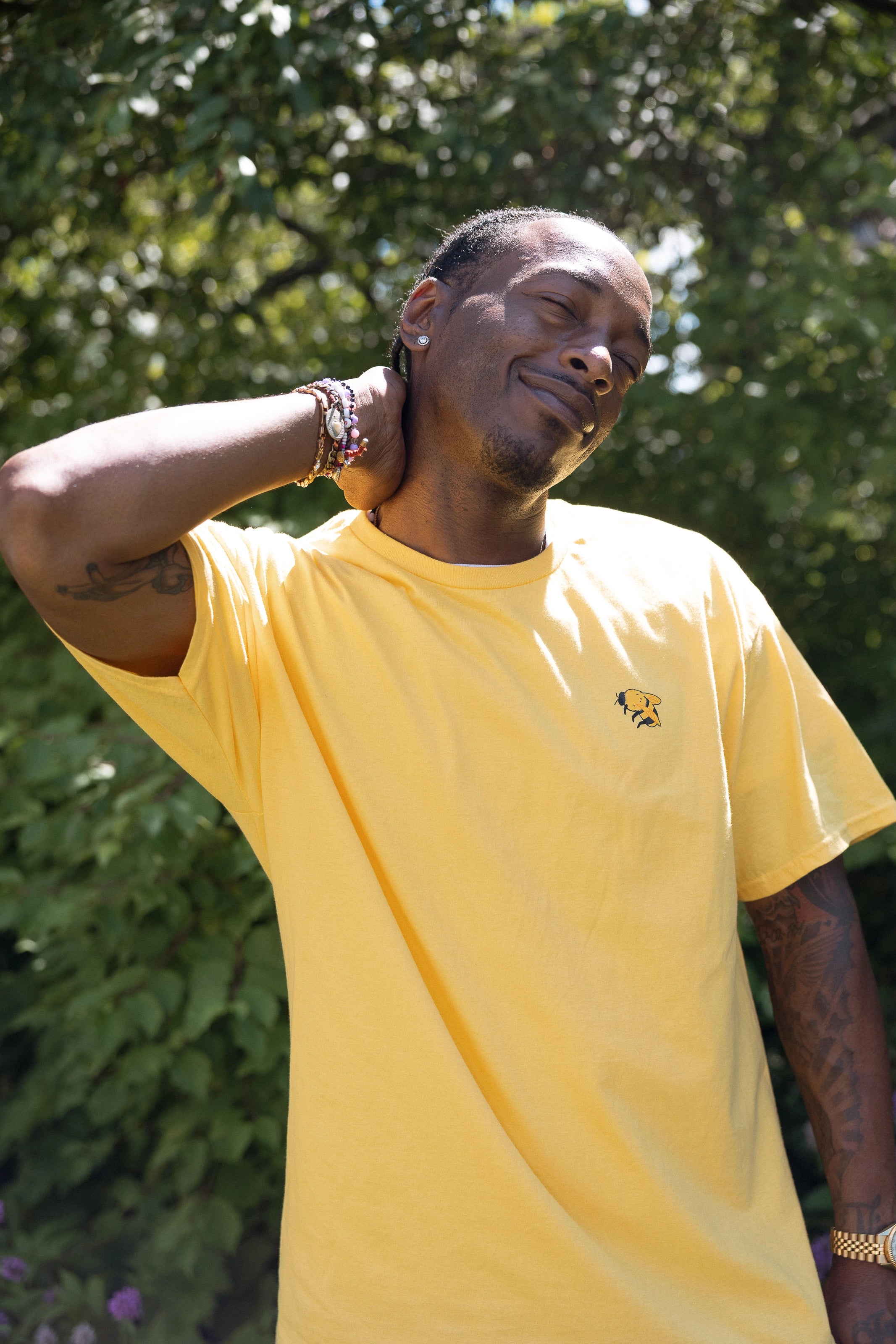 A man wearing a bright yellow crewneck t-shirt with a small bumblebee screenprinted on the left chest, posing outdoors against the foliage of the High Line.