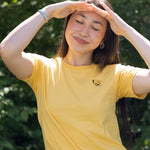 A woman wearing a yellow short-sleeve t-shirt with a small bumblebee printed on the left chest, posing outdoors with her hands shielding her eyes from the sun against a green leafy background.