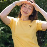 A woman wearing a yellow short-sleeve t-shirt with a small bumblebee printed on the left chest, posing outdoors with her hands shielding her eyes from the sun against a green leafy background.