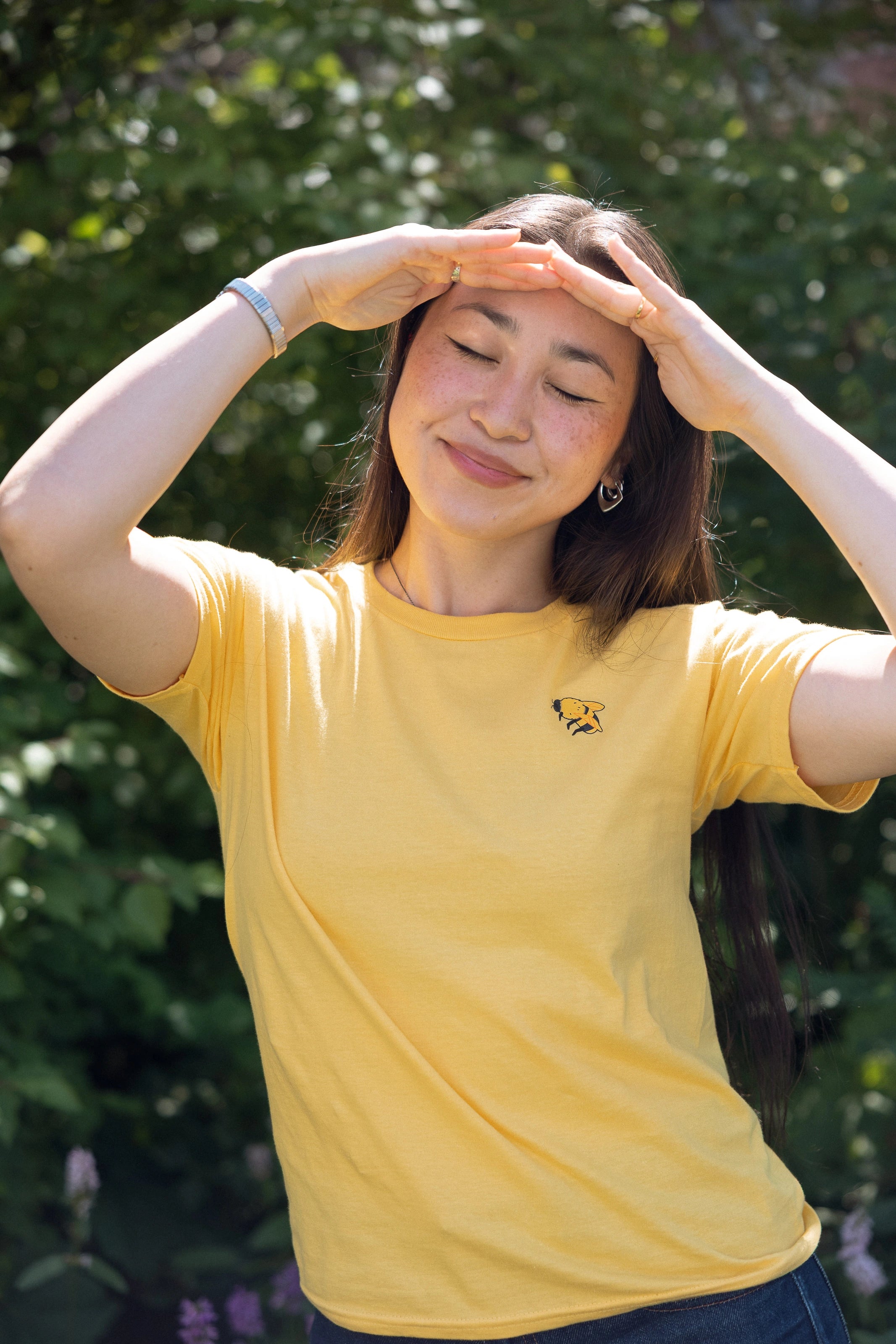 A woman wearing a yellow short-sleeve t-shirt with a small bumblebee printed on the left chest, posing outdoors with her hands shielding her eyes from the sun against a green leafy background.