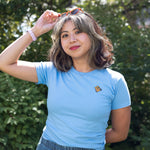Woman wearing a light blue crewneck t-shirt featuring a small Monarch Butterfly on the left chest and denim stands amidst the foliage on the High Line, adjusting her sunglasses.
