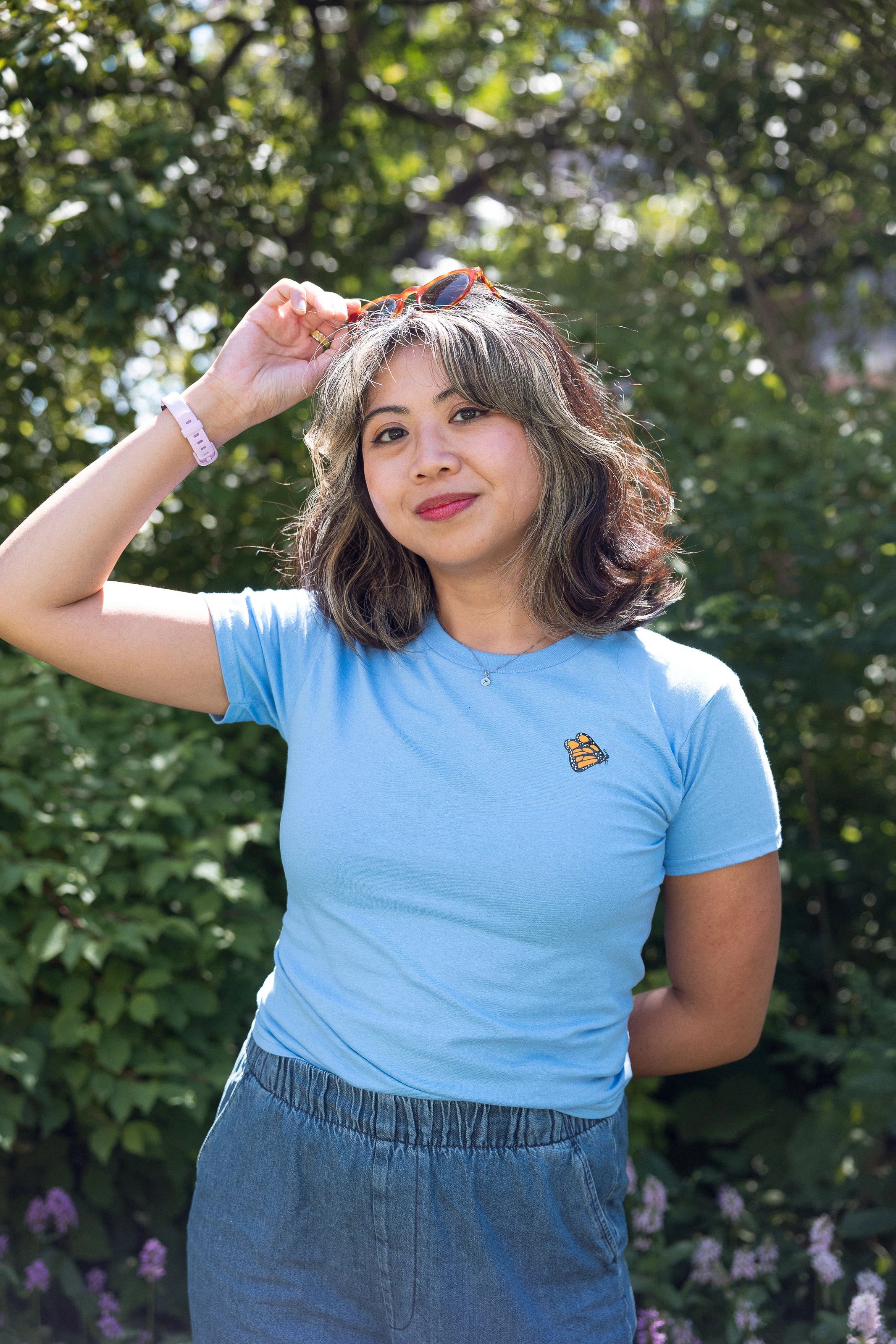 Woman wearing a light blue crewneck t-shirt featuring a small Monarch Butterfly on the left chest and denim stands amidst the foliage on the High Line, adjusting her sunglasses.