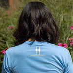 A person viewed from behind wearing a light blue t-shirt with 'protect pollinators' and the High Line icon printed on the back in yellow, amidst the Purple Coneflowers and grasses on the High Line.