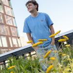 Man wearing a light blue crewneck t-shirt featuring a small Monarch Butterfly on the left chest and light wash jeans stands amidst the wildflowers on the High Line, looking up and smiling. Modern glass buildings and a steel bridge are visible in the background under a clear blue sky.
