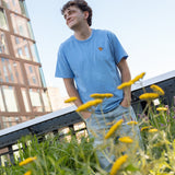 Man wearing a light blue crewneck t-shirt featuring a small Monarch Butterfly on the left chest and light wash jeans stands amidst the wildflowers on the High Line, looking up and smiling. Modern glass buildings and a steel bridge are visible in the background under a clear blue sky.