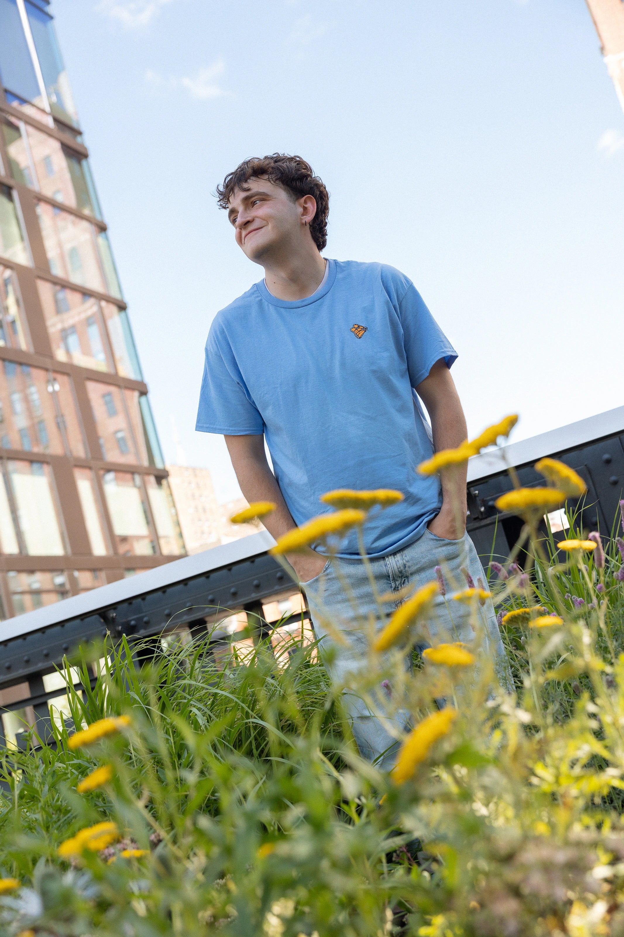 Man wearing a light blue crewneck t-shirt featuring a small Monarch Butterfly on the left chest and light wash jeans stands amidst the wildflowers on the High Line, looking up and smiling. Modern glass buildings and a steel bridge are visible in the background under a clear blue sky.