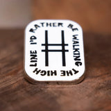 Enamel pin with text 'I'd Rather Be Walking The High Line' resting on wood.