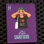 The Sightseer acrylic magnet, featuring a tourist wearing an "I <3 NY" shirt, statue of liberty crown and holding binoculars against a purple background. The magnet is on a black metal locker.