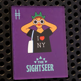 The Sightseer acrylic magnet, featuring a tourist wearing an "I <3 NY" shirt, statue of liberty crown and holding binoculars against a purple background. The magnet is on a black metal locker.