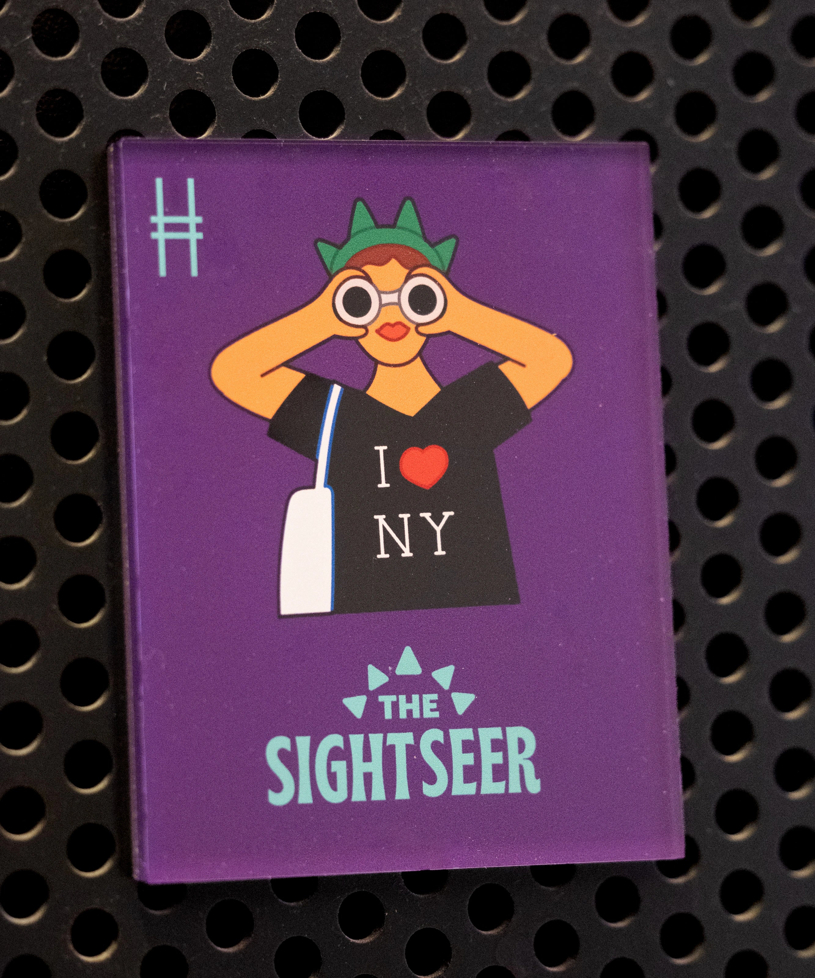 The Sightseer acrylic magnet, featuring a tourist wearing an "I <3 NY" shirt, statue of liberty crown and holding binoculars against a purple background. The magnet is on a black metal locker.