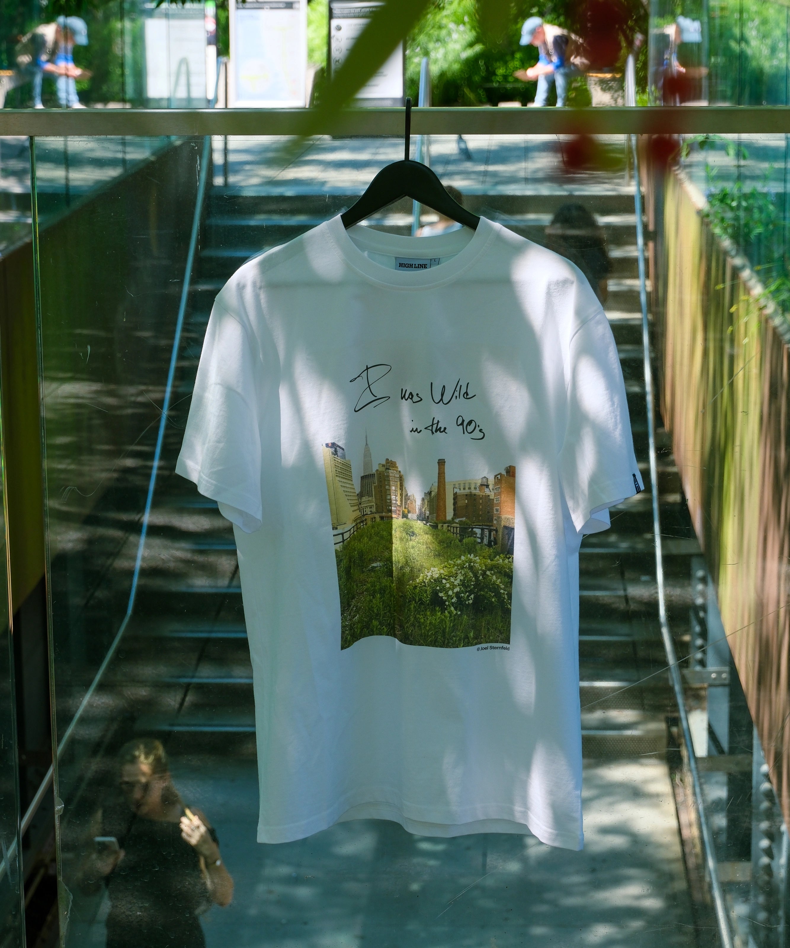 Wild in the 90s Tee hanging on the High Line's Gansevoort Staircase railing. The shirt features one of Joel Sternfeld's iconic photos of the High Line with the phrase "I was wild in the 90s" in his handwriting.