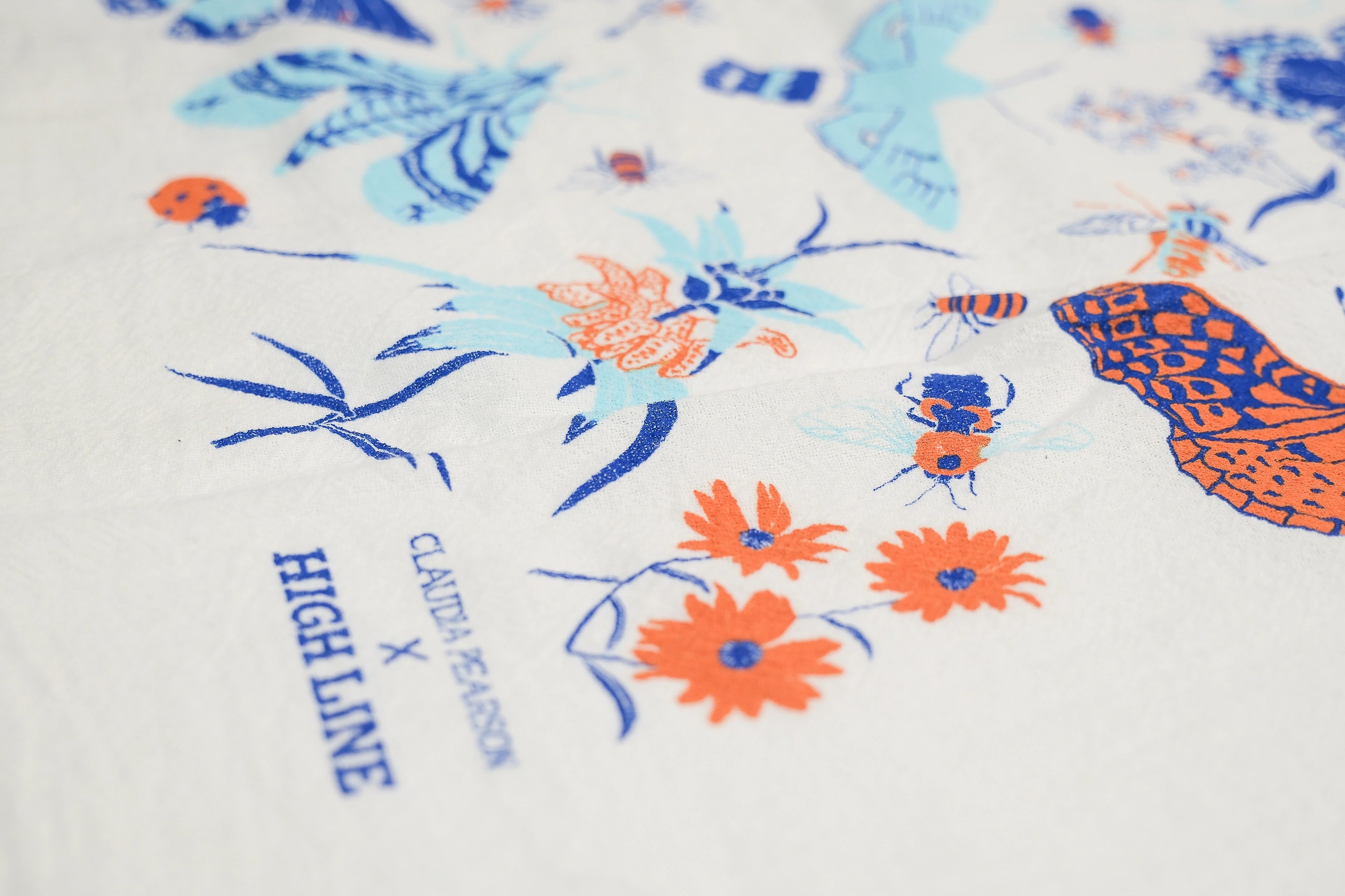 Claudia Pearson x High Line Wildflower Field tea towel, shown in detail. The flour sack cotton tea towel is covered in illustrations of butterflies, ladybugs, moths, other insects and wildflowers in blues and oranges. The logo for the collaboration appears at the bottom.